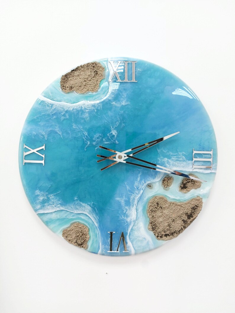 May include: A round blue and white resin clock with Roman numeral markings and silver hands. The clock face features a design of ocean waves and sand.