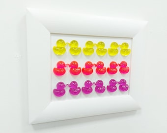 May include: A white frame with three rows of rubber duckies. The top row is yellow, the middle row is red, and the bottom row is purple.