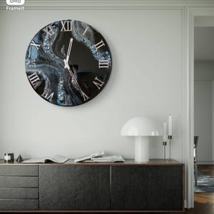 May include: A round wall clock with a black face and Roman numerals. The clock features a unique design with blue and white accents, and is mounted on a white wall. The clock hands are silver.