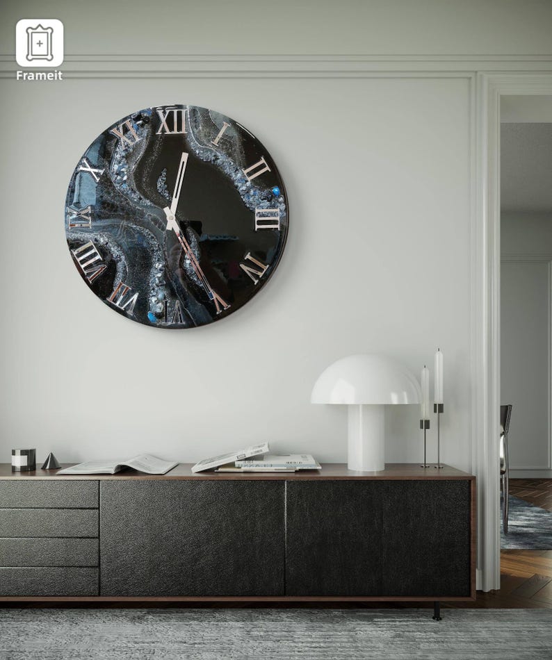 May include: A round wall clock with a black face and Roman numerals. The clock features a unique design with blue and white accents, and is mounted on a white wall. The clock hands are silver.