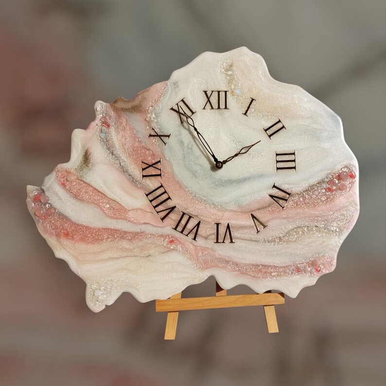 May include: A decorative wall clock with a unique, organic shape. The clock face features Roman numerals and black clock hands. The clock has a white, pink, and beige marbled design, and is supported by a small wooden easel.