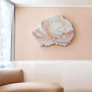May include: A unique wall clock with a white, pink, and gold design. The clock face features gold Roman numerals and hands. The clock has a freeform shape and is mounted on a light pink wall.