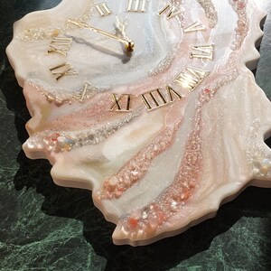 May include: A decorative wall clock with a unique, organic shape. The clock face features gold Roman numerals and hands, set against a white, pink, and clear resin design. The clock measures approximately 12 inches in diameter.