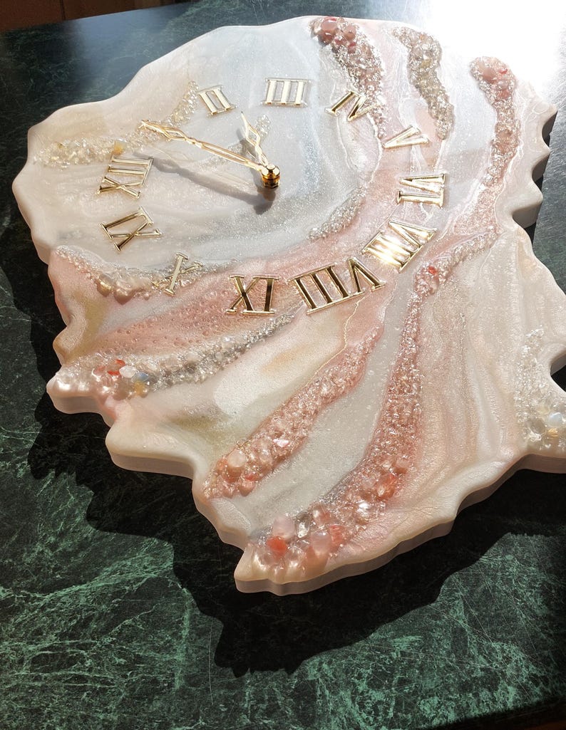 May include: A decorative wall clock with a unique, organic shape. The clock face features gold Roman numerals and hands, set against a white, pink, and clear resin design. The clock measures approximately 12 inches in diameter.