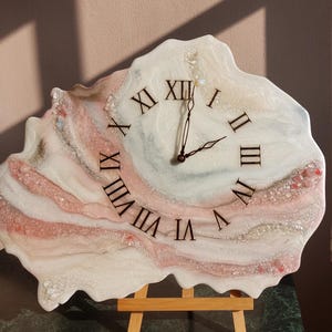 May include: A unique wall clock with a freeform shape, featuring a swirling design in white, pink, and gray tones. The clock face displays black Roman numerals and black clock hands. The clock is mounted on a small wooden easel.