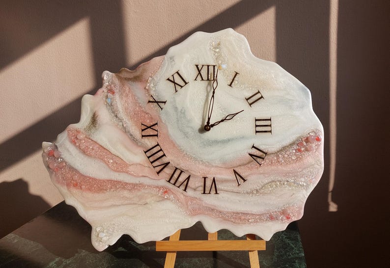 May include: A unique wall clock with a freeform shape, featuring a swirling design in white, pink, and gray tones. The clock face displays black Roman numerals and black clock hands. The clock is mounted on a small wooden easel.
