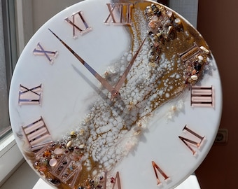 May include: Round wall clock with a white face, featuring rose gold Roman numerals and hands. The clock face has a decorative design with brown and gold accents, and embedded stones. The clock is displayed on a wooden stand.
