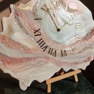 May include: A unique, handmade clock with a resin face. The clock features gold Roman numerals and hands, set against a swirling background of white, pink, and silver. The clock is displayed on a small wooden easel.
