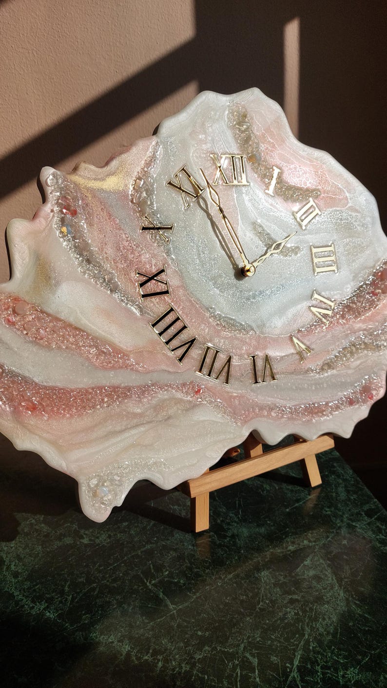 May include: A unique, handmade clock with a resin face. The clock features gold Roman numerals and hands, set against a swirling background of white, pink, and silver. The clock is displayed on a small wooden easel.