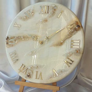 May include: A circular wall clock with a white and beige resin design. The clock face has gold Roman numerals and hands. The clock is displayed on a small wooden easel.