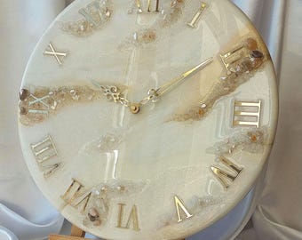 May include: A round wall clock with a white and beige resin design. The clock face features gold Roman numerals and hands. The clock is displayed on a small wooden easel.