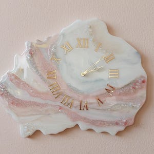 May include: A unique wall clock with a marbled design in white, pink, and silver tones. The clock features gold Roman numerals and hands, creating an elegant and artistic piece. The clock's irregular shape adds to its distinctive appeal.