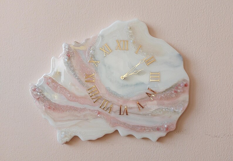 May include: A unique wall clock with a marbled design in white, pink, and silver tones. The clock features gold Roman numerals and hands, creating an elegant and artistic piece. The clock's irregular shape adds to its distinctive appeal.