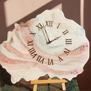 May include: A unique, resin-based wall clock with a freeform shape. The clock face features Roman numerals and black clock hands. The design incorporates shades of white, pink, and gold, with decorative elements.