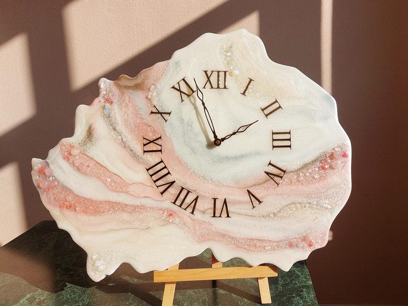 May include: A unique, resin-based wall clock with a freeform shape. The clock face features Roman numerals and black clock hands. The design incorporates shades of white, pink, and gold, with decorative elements.