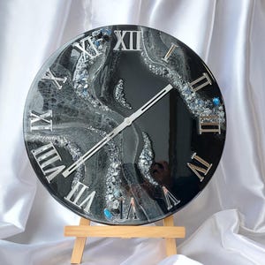 May include: A round black and silver wall clock with Roman numerals. The clock face has a marbled design with silver accents and a wooden easel stand. The clock hands are silver.