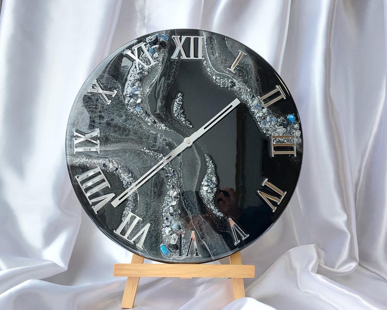 May include: A round black and silver wall clock with Roman numerals. The clock face has a marbled design with silver accents and a wooden easel stand. The clock hands are silver.