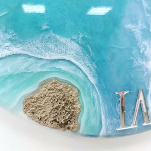 May include: A close-up of a blue and white resin art piece with a sandy brown texture and the letters "LA" in silver.