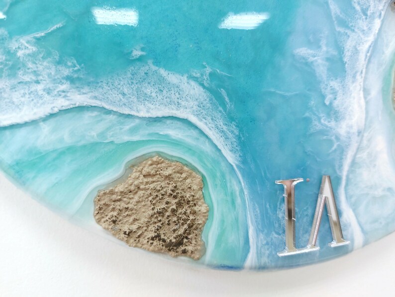 May include: A close-up of a blue and white resin art piece with a sandy brown texture and the letters "LA" in silver.