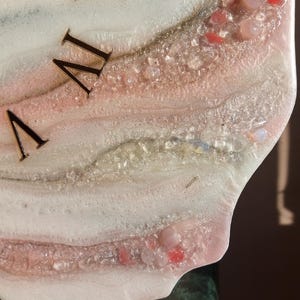 May include: Close-up of a decorative clock face with Roman numerals. The clock face features a white and pink resin design with embedded crystals and gemstones. The numerals are a dark metallic color.