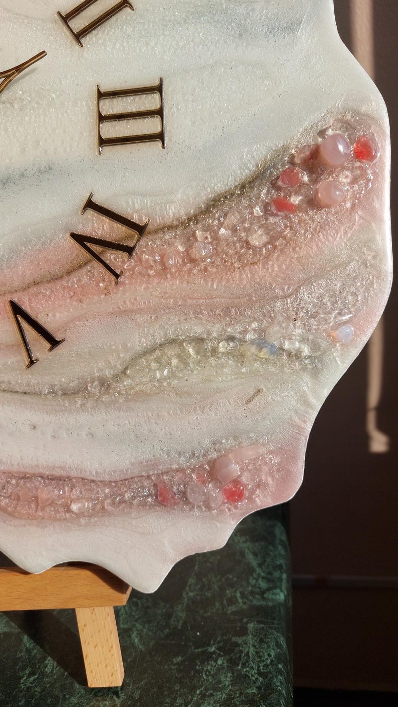 May include: Close-up of a decorative clock face with Roman numerals. The clock face features a white and pink resin design with embedded crystals and gemstones. The numerals are a dark metallic color.