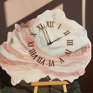 May include: A unique wall clock with a white, pink, and beige resin design. The clock face features black Roman numerals and black clock hands. The clock is displayed on a small wooden easel, showcasing its artistic and decorative nature.
