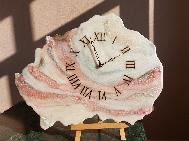 May include: A unique wall clock with a white, pink, and beige resin design. The clock face features black Roman numerals and black clock hands. The clock is displayed on a small wooden easel, showcasing its artistic and decorative nature.