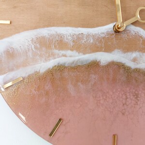 May include: A round clock with a pink and gold resin face. The face features a white wave design with gold accents. The clock hands are gold.