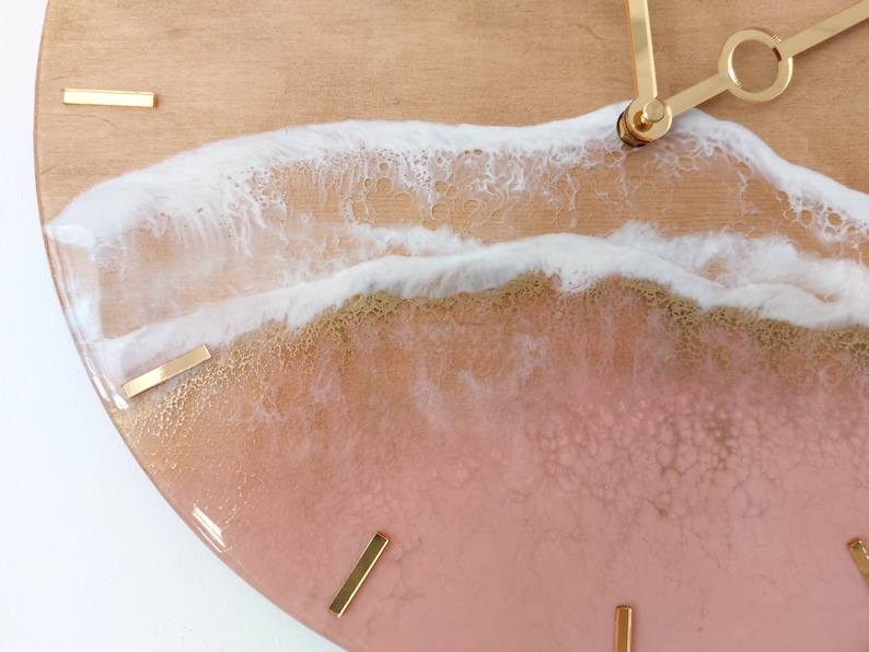 May include: A round clock with a pink and gold resin face. The face features a white wave design with gold accents. The clock hands are gold.