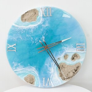 May include: A round blue and white resin clock with Roman numeral markings. The clock face features a design of ocean waves and sand islands.