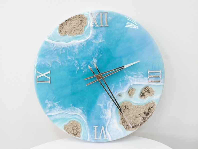 May include: A round blue and white resin clock with Roman numeral markings. The clock face features a design of ocean waves and sand islands.