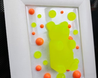 Può includere: Un'opera d'arte incorniciata con una scultura di orso gommoso giallo brillante circondata da punti arancioni e gialli su uno sfondo bianco.