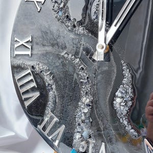 May include: A round clock with a black and gray marbled design, featuring silver Roman numerals and clock hands. The clock face has a textured appearance with embedded stones and a glossy finish. The clock is displayed on a wooden easel.