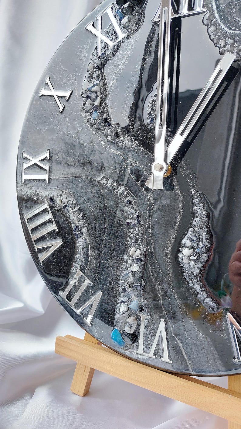 May include: A round clock with a black and gray marbled design, featuring silver Roman numerals and clock hands. The clock face has a textured appearance with embedded stones and a glossy finish. The clock is displayed on a wooden easel.