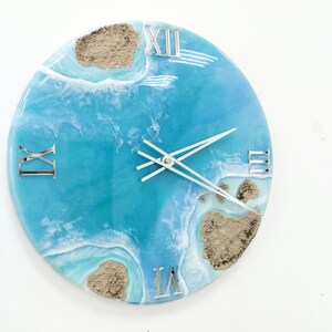 May include: A round blue and white resin clock with Roman numeral markings and a white clock hand. The clock face features a design of ocean waves and sand.