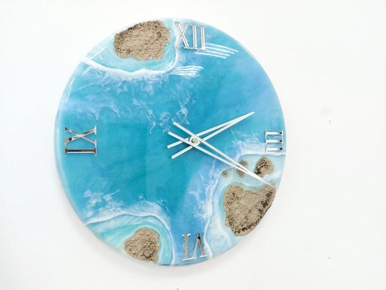 May include: A round blue and white resin clock with Roman numeral markings and a white clock hand. The clock face features a design of ocean waves and sand.
