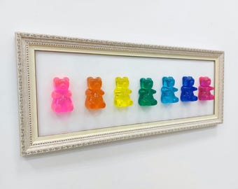 May include: A framed artwork displaying seven gummy bear sculptures in a row. The bears are in a spectrum of colours: pink, orange, yellow, green, blue, and red. The frame is cream-coloured with decorative detailing.