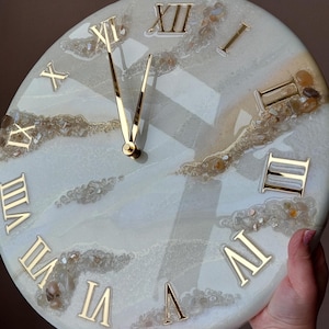 Op de afbeelding: Een ronde wandklok met een wit en lichtblauw marmerontwerp. De klok heeft gouden Romeinse cijfers en gouden wijzers. De wijzerplaat heeft een unieke, getextureerde uitstraling met ingebedde stenen en een glanzende afwerking.