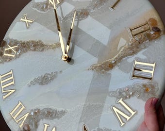 May include: A round wall clock with a white and light blue marbled design. The clock features gold Roman numerals and gold hands. The clock face has a unique, textured appearance with embedded stones and a glossy finish.