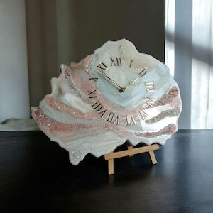 May include: A unique, handcrafted clock with a seashell design. The clock face features Roman numerals and gold-colored hands. The clock is supported by a small wooden easel and has a color palette of white, pink, and silver.