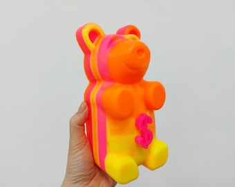 Può includere: Un salvadanaio a forma di orso gommoso di colore arancione, giallo e rosa acceso con un simbolo del dollaro rosa sul davanti.