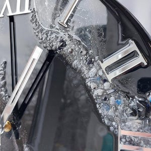 May include: Close-up of a black clock with silver Roman numerals and hands. The clock face has a textured, resin-like appearance with embedded stones in shades of gray and blue. The clock's design is modern and decorative.