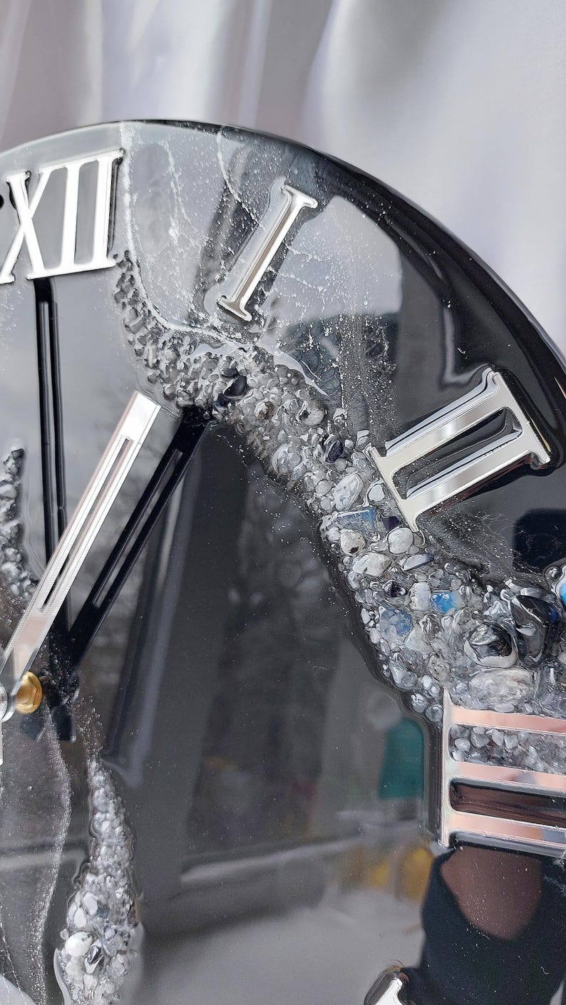 May include: Close-up of a black clock with silver Roman numerals and hands. The clock face has a textured, resin-like appearance with embedded stones in shades of gray and blue. The clock's design is modern and decorative.