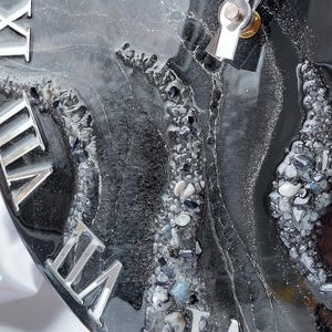May include: Close-up of a round clock with a black and gray resin surface, embedded with small stones and silver Roman numerals. The clock hands are silver. The clock is on a wooden stand.