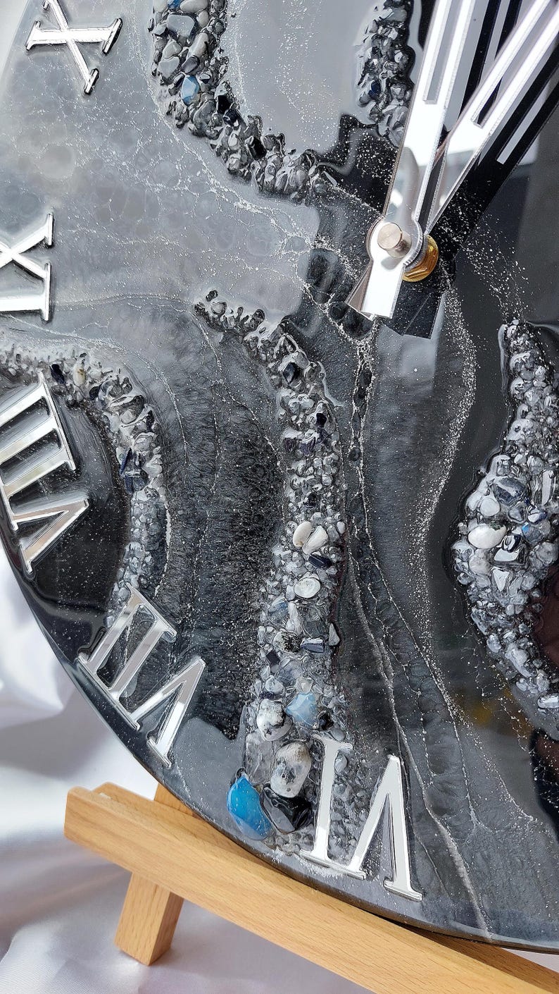 May include: Close-up of a round clock with a black and gray resin surface, embedded with small stones and silver Roman numerals. The clock hands are silver. The clock is on a wooden stand.