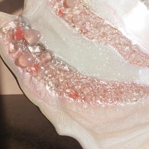May include: Close-up of a decorative resin piece with a unique, organic shape. The piece features a blend of white, pink, and clear resin, embedded with small, pink and clear stones. Gold flecks add a touch of shimmer.