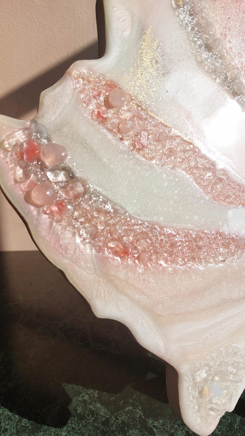 May include: Close-up of a decorative resin piece with a unique, organic shape. The piece features a blend of white, pink, and clear resin, embedded with small, pink and clear stones. Gold flecks add a touch of shimmer.