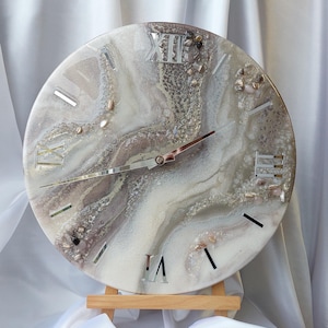 Può includere: Orologio da parete rotondo con un design marmorizzato nei toni del bianco, grigio e marrone. Il quadrante dell'orologio presenta numeri romani e lancette argentate. L'orologio è esposto su un piccolo cavalletto di legno.