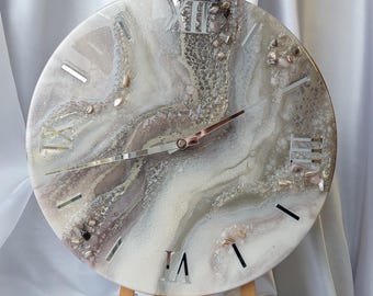 Puede incluir: Reloj de pared redondo con diseño veteado en tonos blancos, grises y marrones. La esfera del reloj presenta números romanos y manecillas plateadas. El reloj se exhibe sobre un caballete de madera.