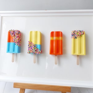 May include: A framed artwork featuring four colorful popsicle sculptures. Each popsicle is a different color, including blue, yellow, and orange, and some are decorated with sprinkles. The popsicles are mounted on a white background.